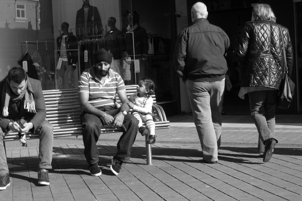 A man in a striped polo and a small girl sit on a steel bench while another man looks at his phone