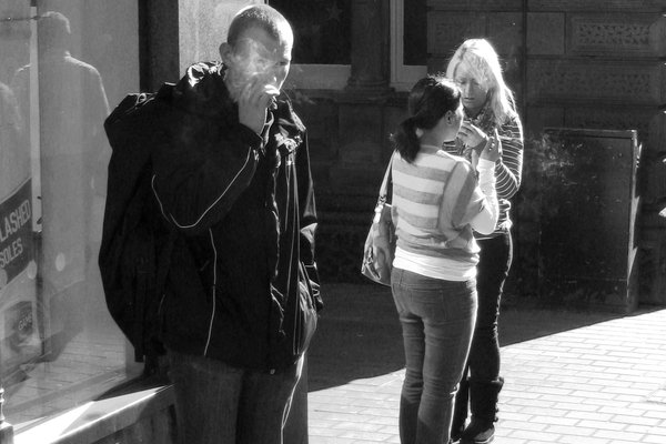 A man breathes out cigarette smoke in the sun while two women share a light behind him