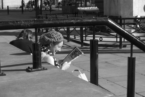 A woman in a knitted beret reads a paper, seen between railings and a stone ledge