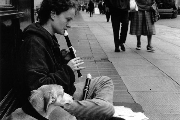A young woman plays a recorder on the pavement with a dog beside her as passers-by walk on
