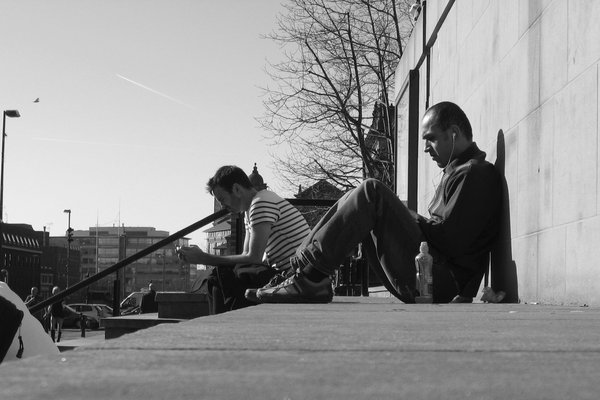 Two men sit on a sunlit ledge, one looking at his phone and one leaning back with earphones in
