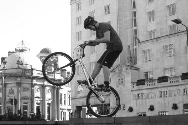 A trials rider balances on the edge of a wall with his rear wheel raised and a broad smile