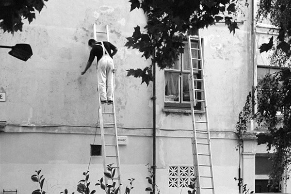 A painter in white overalls stands on a ladder and touches a weathered wall